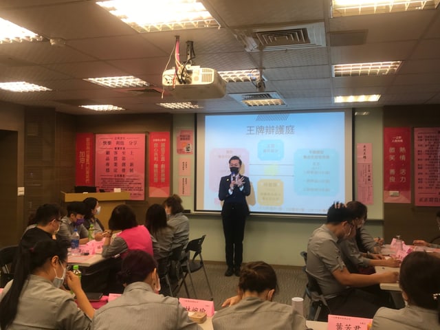 在企業太學課堂上以「王牌辯護庭」辯論形式帶課：投影幕是「開發 vs 不應開發集合式住宅市場」的攻防架構，學員在座位上持桌牌參與